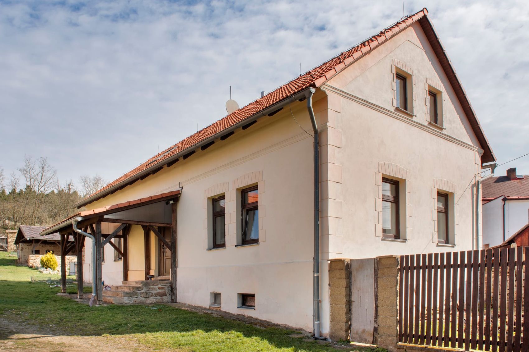 Světlá chalupa s oranžovou střechou, dřevěným plotem a zahradou za vsí